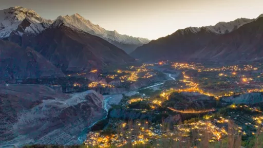 A wide valley surrounded by tall, snow-capped mountains is illuminated by clusters of warm lights from villages below at dusk. A river winds through the center of the valley, reflecting the fading light of the sky.