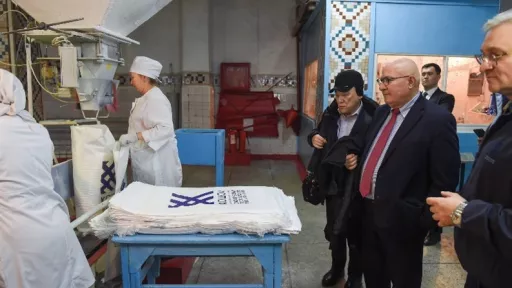 In a factory, workers in white uniforms handle sacks at a machine while people in business attire observe. Stacks of printed white sacks are on a blue table nearby.