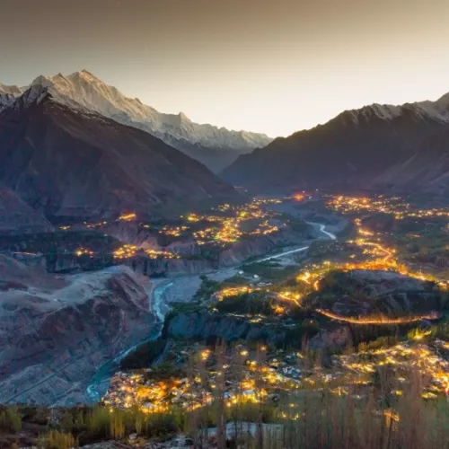 A wide valley surrounded by tall, snow-capped mountains is illuminated by clusters of warm lights from villages below at dusk. A river winds through the center of the valley, reflecting the fading light of the sky.
