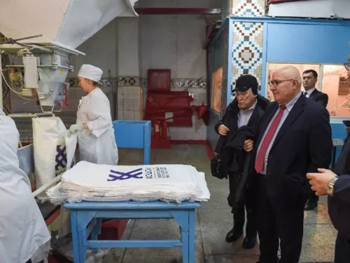In a factory, workers in white uniforms handle sacks at a machine while people in business attire observe. Stacks of printed white sacks are on a blue table nearby.